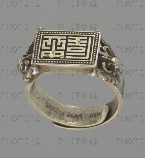 Ring, Chinese, Chinese art, Qing 1644–1911, body ornament, jewelry, hand ornament, bats iconography, Shield with ideographic signs supported by two bats, 19th century, China, jewelry, diameter 2 cm, A metal ring shown against a gray background with a rectangular top plate featuring a raised geometric pattern framed by a beaded border; the band and decorative elements appear aged with a patina, showing a mix of dull gold and darker tarnished tones, and there are sculpted ornamental shapes on the sides of the top plate resembling curled forms in similar gold and dark hues.