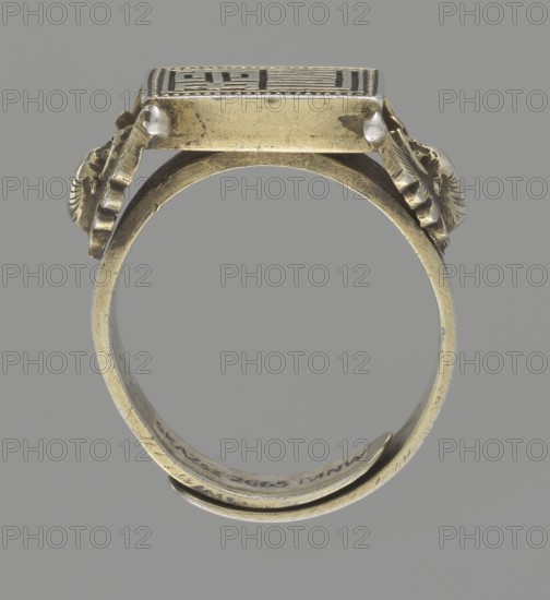 Ring, gift, Chinese, Chinese art, Jagniatkowska, Magdalena 1866-1944, Jagniatkowski, Wladyslaw 1856-1930, Qing 1644–1911, body ornaments, jewelry, hand ornaments, bats iconography, Shield with ideographic signs supported by two bats, 19th century, China, jewelry, dia. 2 cm, A single metal ring shown from the side against a neutral gray background, the ring appearing to be made of aged yellowish metal with areas of darker tarnish and lighter reflections, featuring an elevated rectangular top with decorative relief and small rounded protrusions at its corners; the band curves into a near-circle with a slightly flattened underside and shows surface scratches and wear, while ornamental elements resembling small folded or scalloped shapes flank the top on both sides in similar yellowish metal with darker shadowed crevices.