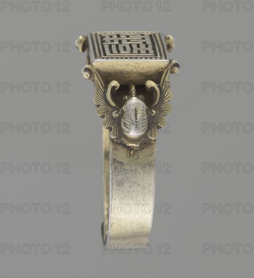 Ring, Chinese, Qing 1644–1911, 19th century. Gold jewelry hand ornament, diameter 2 cm. Design: shield with ideographic signs supported by two bats; bats iconography. ., A close-up of a metallic bracelet or cuff shown vertically against a plain gray background, featuring a decorative top section with a rectangular raised element and an ornate insect or scarab motif beneath it; the metal shows a mix of gold and silver tones with areas of patina and darker tarnish, and the decorative wings and curved details are highlighted in a warmer golden color while the band displays a more muted silvery finish.