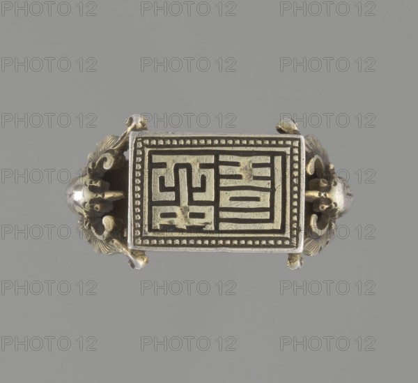 Ring, Chinese, Qing 1644–1911, 19th century. Jewelry; hand ornament. Shield with ideographic signs supported by two bats motif; bats iconography. Diameter 2 cm. ., A rectangular metallic object with ornate curved sides rests against a plain gray background; the rectangle has a raised border with a dotted inner edge and an inset panel featuring a pattern of raised linear shapes, and the curved side elements include decorative scroll-like and floral motifs with rounded bulbous forms. The visible colors are metallic gold, silver, and gray.