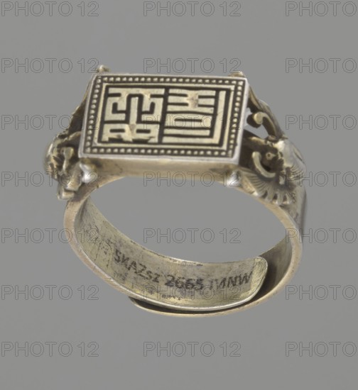 Ring, Chinese, Chinese art, Qing 1644–1911, body ornament/jewelry/hand ornament, motif: shield with ideographic signs supported by two bats, 19th century, China, diameter 2 cm, A metal ring photographed against a plain gray background, showing a rectangular top with a raised patterned design and a dotted border, ornate sculpted sides with decorative motifs, and a curved band with visible surface wear; the visible colors are silver, gray, and muted gold.