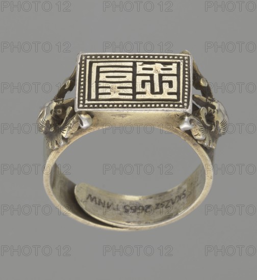 Ring, gift, Chinese, Chinese art, Qing 1644–1911, body ornament, jewelry, hand ornament, bats iconography, Shield with ideographic signs supported by two bats, 19th century, China, diameter 2 cm, A metallic ring photographed against a plain gray background, featuring a rectangular top plate with a raised patterned design and a dotted border; the ring body and decorative elements are primarily gold-toned with some darker tarnished areas and small silver-toned accents, and a few sculpted ornamental motifs on the shoulders resembling stylized animal heads; the inside of the band shows signs of wear and darker spots.
