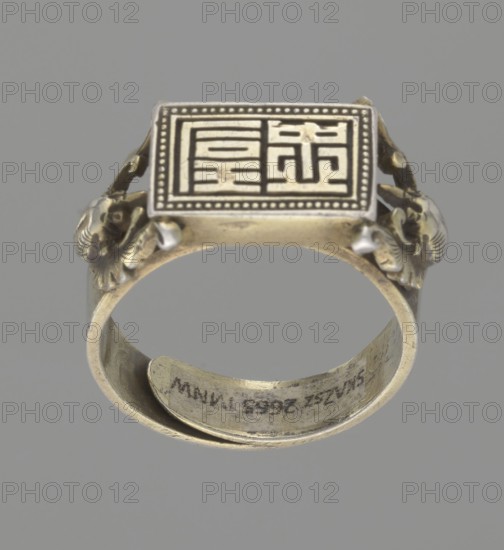Ring, Chinese, Chinese art, Jagniatkowska, Magdalena 1866–1944, Jagniatkowski, Wladyslaw 1856–1930, Qing 1644–1911, body ornaments, jewelry, hand ornaments, bats iconography, Shield with ideographic signs supported by two bats, 19th century, China, jewelry, diameter 2 cm, A metallic ring shown against a gray background with a rectangular top plate featuring a raised geometric maze-like pattern and a dotted border; the ring band has decorative sculpted elements on the sides resembling floral or animal heads and a curled inner extension; visible colors include silver, gold-toned metal, dark gray accents within the pattern, and the neutral gray background.