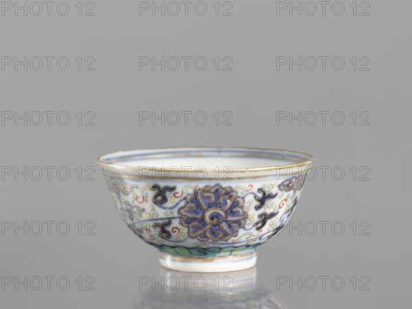 Bowl, Chinese, Qing dynasty 1644–1911, 19th century; colored porcelain vessel with plant and floral ornaments including lotuses and lotus tendril motif, floral motifs on the reverse of the rim; diameter 6.8 cm., A small ceramic bowl sits on a reflective surface against a neutral gray background, showing a rounded rim and a short foot; the bowl is predominantly white with decorative motifs in blue and purple, accented with green, yellow, red, and black, and the rim and foot edges have a thin gold or brass-colored band; the exterior displays a repeating floral or scroll-like pattern in deep blue and purple with sinuous outlines and small accent swirls in red and yellow, and there is a green band near the foot.