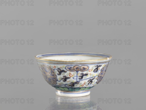 Bowl, Chinese, Qing dynasty 1644–1911, 19th century. Ceramic colored porcelain vessel with plant and floral ornaments—flowers, lotuses, lotus tendril motif, floral motifs on the reverse of the rim. Diameter 6.8 cm., A small ceramic bowl with a rounded shape and a short foot sits against a plain gray background, showing a white interior rimmed with a thin band of gold and a blue line, and an exterior decorated with intricate painted floral and scroll motifs in blue, dark blue, green, yellow, red, and gold accents, with a glossy finish that produces a faint reflection beneath it.