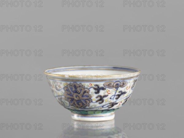 Bowl, 19th century, China; Qing dynasty 1644–1911; colored porcelain vessel with plant and floral ornaments including lotuses and lotus tendril motifs, floral motifs on the reverse of the rim; diameter 6.8 cm., A small ceramic bowl sits on a reflective surface against a plain gray background; the bowl is primarily white with decorative patterns in blue, gold, green, and red, including a prominent blue floral motif outlined in gold and several scrolling cloud-like designs, a thin gold rim at the top, and a green band near the base, with the interior appearing white and the exterior showing glazed sheen.