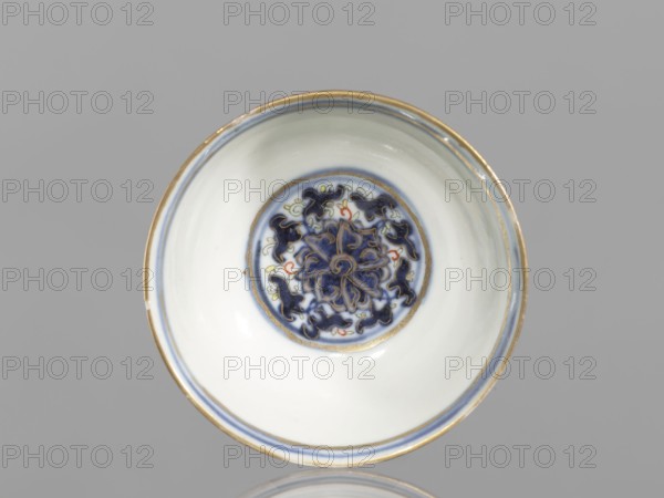 Bowl, 19th century, China; Qing dynasty 1644–1911; colored porcelain vessel, diameter 6.8 cm; decorated with plant and floral ornaments—flowers, lotuses, and lotus tendril motif—with floral motifs on the reverse of the rim., A shallow round bowl viewed from above with a glossy white interior and a decorative circular medallion at the center featuring a floral-like pattern in dark blue with small accents of red and pale yellow, surrounded by concentric bands in blue and gold; the rim has a thin gold edge and a thin blue line just inside it, and the background is a uniform light gray.