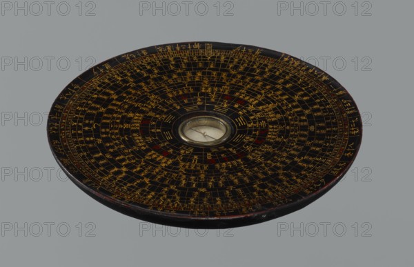 Feng shui compass luopan, Chinese, Qing 1644–1911, 19th century. San yuan luo pan also known as yi pan or jiang pan with the ring for the 64 hexagrams of the I-Ching and a 24-direction ring; used by practitioners of the Three Cycles System of Feng Shui including Flying Stars Time Space. Function: navigational/astrological measuring instrument. Diameter 17.0 cm., A circular, flat object with concentric rings of fine markings and grid-like divisions across its dark surface, featuring a central round glass-covered element with a small needle; the surface shows a pattern of primarily gold lines and symbols with occasional red accents on a black background, and the rim has a worn dark edge with hints of brown; the overall background is a uniform gray.