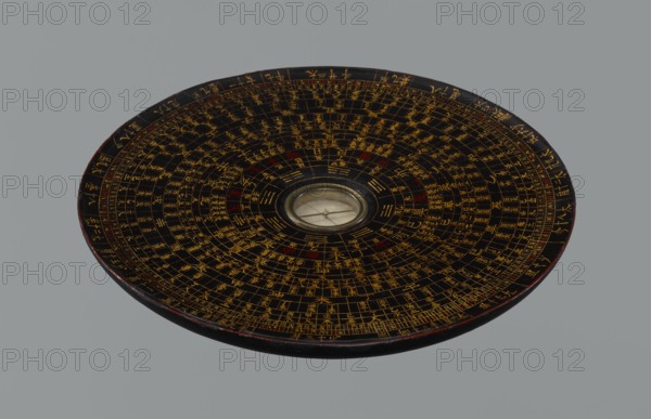Feng shui compass luopan, Chinese, Qing 1644–1911, 19th century. San Yuan Luo Pan also known as yi pan or jiang pan containing the ring for the 64 hexagrams of the I Ching and a 24-direction ring. Used by practitioners of the Three Cycles System of Feng Shui including Flying Stars Time Space. Material: measuring instrument; diameter 17.0 cm. Subjects: navigational objects, astrology, calendars, Chinese Zodiac., A circular lacquered object with a central glass-covered circular inset, displaying a concentric pattern of small rectangular segments radiating outward; the surface is primarily black with intricate markings in gold and some accents in red, and the central inset shows a pale, reflective area beneath glass. The object has a slightly raised rim with subtle wear revealing a dark reddish edge, and it rests against a uniform gray background.