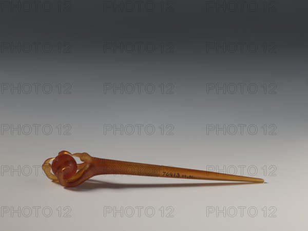 Hairpin, gift, repossession, Chinese art, Chinese culture, head ornament, women's ornament, toiletry, Qing 1644–1911, transparent amber, 17th–18th century, China, jewelry, length 13.6 cm, A slender object in shades of amber and brown rests horizontally on a smooth, light surface, tapering to a pointed end; the wider end features a rounded, glossy bead held by curved, claw-like prongs with textured surfaces, and the shaft displays a repeating crosshatched texture; the background transitions from light near the surface to dark gray toward the top, and there is a soft shadow beneath the object.