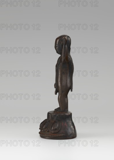Young Buddha, unknown sculptor; Buddha iconography, figurine on lotus throne; Chinese Buddhist art, Qing dynasty 1644–1911, 19th/20th century, China; sculpture; height 16.7 cm; children., A small dark brown carved figure standing on a rounded decorative base, shown in profile facing left, with a smooth elongated head and simplified facial features, one arm raised with the hand near the head and the other arm hanging down, the body and limbs are slender and unadorned, the base has carved petal-like motifs and slight red traces on the dark surface, the background is a gradient of pale gray to off-white.