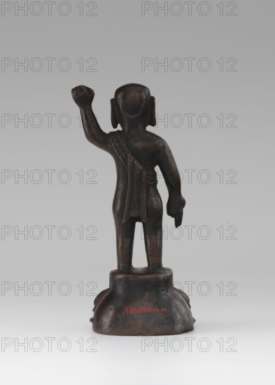 Young Buddha, unknown sculptor, gift, repossession Krakow, Buddha iconography, figurine on lotus throne, Chinese art, Chinese culture, Chinese Buddhism, children, Qing 1644–1911, Buddhist art, 19th/20th century, China, sculpture, height 16.7 cm, A small dark brown statue viewed from behind standing on a rounded base, with a raised arm and a lowered arm, a draped cloth over one shoulder and down the back, short legs and rounded head, and visible colors including dark brown, hints of black, and a neutral gray background.