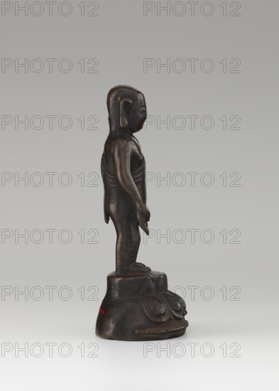 Young Buddha; unknown sculptor; gift, repossession; Buddha iconography; figurine on lotus throne; Chinese art; Chinese Buddhism; children; Qing dynasty 1644–1911; Buddhist art; 19th–20th century; China; sculpture; height 16.7 cm, A dark brown, patinated figurine of a standing human-like figure shown in profile on a rounded base, with smooth limbs and a stylized head, a few subtle highlights of lighter brown where the surface is worn, and a neutral gray background.