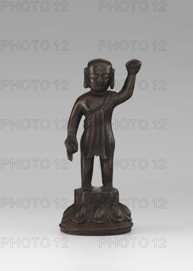 Young Buddha, unknown sculptor, gift, repossession Krakow, Buddha iconography, figurines, lotus throne iconography, Chinese art, Chinese culture, Chinese Buddhism, children, Qing 1644–1911, Buddhist art, 19th/20th century, China, sculpture, height 16.7 cm, A small dark brown metal statue of a standing figure on a round decorated base is visible; the figure has a raised right arm and a lowered left arm with a hand gesture, a short garment draped over one shoulder, stylized hair or head covering, and a smooth patina showing darker and lighter brown tones, set against a neutral gray background.