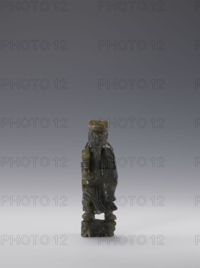 Sage, unknown sculptor, figurine, Chinese culture, Chinese art, sages, Qing dynasty 1644–1911, grey-green soapstone, 19th century, China, sculpture; height 13.8 cm, width 4.4 cm, depth 3.1 cm, A small carved figurine stands on a carved base, the figure wearing a long flowing robe and a tall hat, holding a rounded object in one hand and a staff or similar object in the other; the carving shows facial features and a long beard, with draped sleeves and detailed folds in the clothing. The object is primarily shades of dark green and olive with some lighter greenish-yellow areas and slight mottling, set against a neutral gradient background of gray transitioning to a pale gray near the bottom.