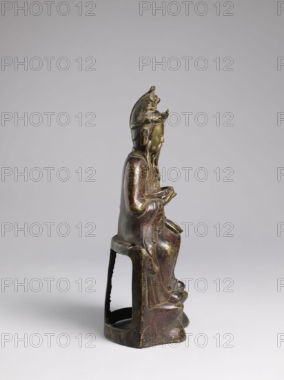 Mythical emperor, Chinese sculptural figurine; Qing dynasty 1644–1911, 19th century; material/technique: sculpture; subject: mythical emperor wearing dragon robes with dragon iconography; culture: Chinese; height 41.0 cm., A seated bronze figure shown in profile on a small open-backed stool, with a long beard and a tall headdress topped by a small animal-like form; the figure wears flowing robes with etched decorative patterns and has hands held in front as if holding something; colors visible include dark brown, greenish-gold, and hints of reddish-brown on the metal surface, with areas of patina and wear against a light gray background.