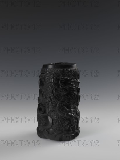 Vase, Chinese, Qing dynasty 1644–1911, 19th century. Vessel with bas-relief decoration depicting dragons. Height 24.5 cm, diameter 14.3 cm., A tall cylindrical carved object sits centered on a smooth surface, rendered in deep black with a slight sheen; the background graduates from dark gray at the top to lighter gray near the surface. The cylinder's exterior shows an intricate raised relief with flowing, curving shapes and textured rough areas, including spiral motifs and wave-like forms around the base and sides. The top is flat and dark, contrasting subtly with the slightly lighter highlights on the carved edges. Overall the visible colors are black and various shades of gray.