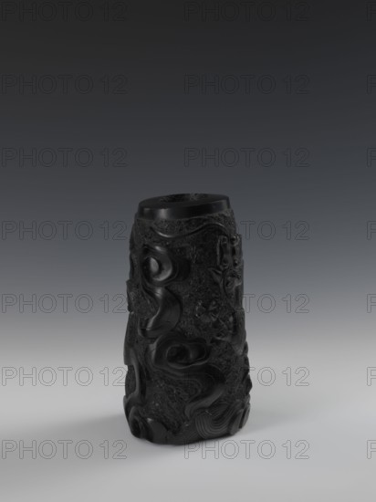 Vase, gift, Chinese art; Chinese; Qing dynasty 1644–1911; 19th century. Vessel with bas-relief decoration of dragons. Height 24.5 cm; diameter 14.3 cm., A single cylindrical carved object stands upright on a smooth surface, appearing dark black with a glossy sheen and matte textured areas; its surface is covered in raised swirling and organic relief patterns that form flowing curves and floral motifs, with areas of rough stippled texture contrasting with the polished carved sections; the top rim is visible and flat, and the base sits casting a soft shadow; the background shows a gradient from dark gray at the top to lighter gray near the bottom.