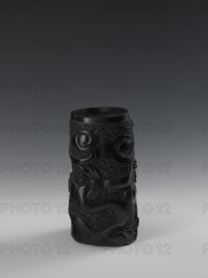Vase, Chinese, Qing dynasty 1644–1911, 19th century. Vessel with bas-relief decoration of dragons. Height 24.5 cm; diameter 14.3 cm., A cylindrical black carved object stands upright on a smooth light-gray surface against a dark gray gradient background; the object has a textured body with glossy carved raised motifs that swirl and curl around it, including serpentine and rounded forms, and both rough matte and polished shiny black areas creating contrasting highlights and shadows.