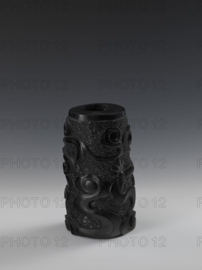 Vase, Chinese, Qing dynasty 1644–1911, 19th century. Bas-relief decoration with dragons iconography. Vessel, height 24.5 cm, diameter 14.3 cm., A dark cylindrical carved object stands upright on a smooth surface, showing a textured body with raised swirling and curving motifs that wrap around it; the material appears glossy in places and more matte and rough in others, with visible variations in surface texture. The top has a recessed circular opening and a smoother rim. The lighting casts soft shadows beneath the object. The visible colors in the image are black and shades of gray.