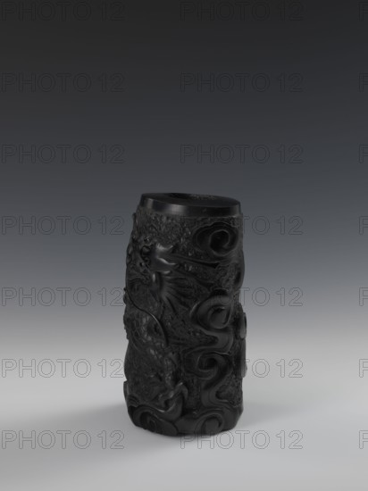 Vase, Chinese, Qing dynasty 1644–1911, 19th century. Vessel with bas-relief decoration of dragons. Height 24.5 cm; diameter 14.3 cm., A dark cylindrical carved object stands upright on a light surface against a gradient background; the object is predominantly black with subtle highlights and a slightly glossy finish, and its surface features a carved design showing flowing, curved shapes and textured areas with raised relief that suggest organic motifs; the surrounding colors include shades of gray in the surface and background, and black on the object itself.