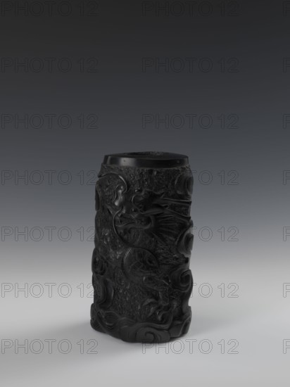 Vase, gift, Chinese art, Chinese, Qing 1644–1911, dragons iconography, bas-relief decoration, 19th century, China, vessel, height 24.5 cm, diameter 14.3 cm, A dark cylindrical carved object stands upright on a smooth surface, its surface featuring raised swirling and curved patterns and a prominent carved dragon motif wrapping around the body; the object is predominantly black with subtle highlights that reveal textured rough areas and polished smooth sections, and it casts a soft shadow on the light gray surface beneath it against a gradient background that transitions from light gray near the base to dark gray toward the top.