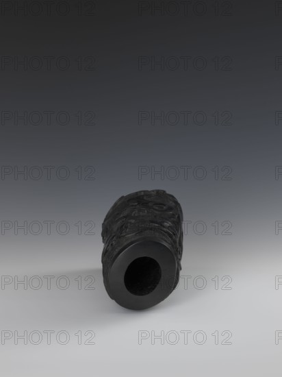 Vase, Chinese, Qing dynasty 1644–1911, 19th century. Porcelain vessel with bas-relief dragon decoration. Height 24.5 cm; diameter 14.3 cm., A cylindrical black object lies on a smooth light gray surface against a gradient dark gray background, showing a hollow circular opening facing the viewer and textured carved or raised patterns along its curved exterior; visible colors include black, dark gray, and light gray.