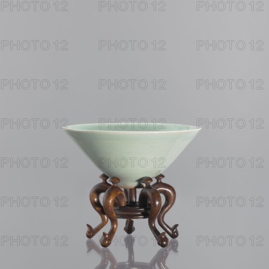 Bowl on base; maker, Shallow, round, profiled base with a round hole in the center; on five legs—stylized geese with bent necks protruding above the base. Chinese; Qing dynasty 1644–1911, 18th–19th century. Ceramic vessel, celadon gray-green glaze. Height 6.1 cm; diameter 12 cm., A pale green ceramic bowl with a smooth glazed surface sits on a small dark brown wooden stand; the bowl has a wide flaring rim and a rounded interior, and the stand features a group of curving, carved legs with rounded ends and a circular central support, all shown against a neutral light gray background with reflections visible beneath the stand.