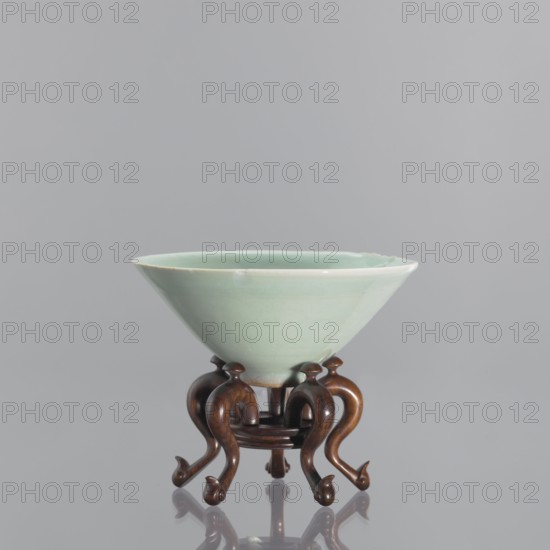 Bowl on base, gift; Chinese art; Chinese, Qing 1644–1911; vessel, ceramics, celadon, gray-green glaze celadon. Shallow, round, profiled base with a round hole in the center; on five legs—stylized geese with bent necks protruding above the base. 18th/19th century, China; height 6.1 cm, diameter 12 cm., A pale celadon-green bowl with a smooth glazed surface sits on an ornate wooden stand; the bowl has a wide flared rim and a shallow, rounded interior, and the glaze shows subtle variations in the green tone. The stand is a dark brown wood with a polished finish and a sculptural, curved design featuring a group of several sinuous legs that curl at the ends, creating small rounded feet; the wood displays warm brown and reddish-brown hues. The background is a uniform light gray, and the bowl and stand cast soft reflections on a glossy surface beneath them.