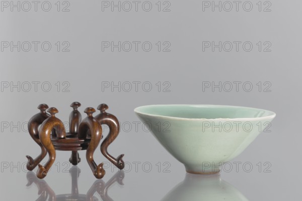Bowl on base, maker, Shallow, round, profiled base with a round hole in the center; mounted on five legs formed as stylized geese with bent necks protruding above the base. Chinese celadon ceramic vessel, gray-green glaze; Qing dynasty 1644–1911, 18th–19th century. Height 6.1 cm; diameter 12 cm., A small wooden stand with a warm brown finish and curved, ornate legs sits next to a pale green glazed ceramic bowl; the stand has a glossy surface and rounded top elements, and the bowl has a smooth, slightly flared rim and a soft, matte-to-gloss gradient on its exterior, with both objects reflected on a shiny gray surface and set against a neutral light gray background, showing colors including brown, pale green, gray, and hints of white.