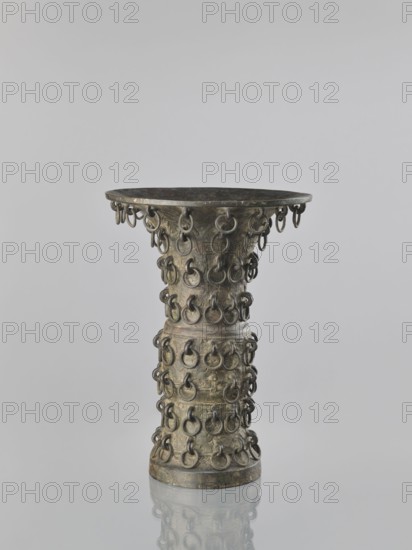 Zun-type vase, Chinese, 19th century Qing dynasty, 1644–1911, ceremonial/ritual vessel with taotie iconography, height 33.0 cm, diameter 23.8 cm., A tall, hourglass-shaped metal object with a flared top and a round base sits against a plain light gray background, its surface showing a weathered patina in shades of dark bronze, greenish-gray, and muted brown; the body is covered with a pattern of several small metal rings attached in rows, and a few rings hang from the rim of the top; the object casts a soft reflection on the glossy surface beneath it.