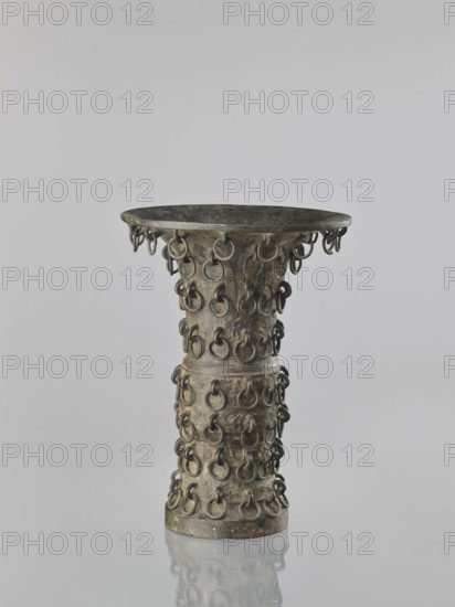 Zun-type vase, Chinese, 19th century Qing 1644–1911, ceremonial/ritual vessel, taotie iconography, vessel, height 33.0 cm, diameter 23.8 cm, A tall, hourglass-shaped metal vessel with a wide, shallow rim and a narrow waist is shown standing on a reflective surface; its surface is studded all around with a repeating pattern of small metal rings attached in rows, and the metal shows a patina with visible greenish and brownish tones along with darker gray and black areas.