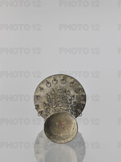 Zun-type vase, Chinese, 19th century Qing dynasty, 1644–1911, ceremonial/ritual vessel with taotie iconography, height 33.0 cm, diameter 23.8 cm., An aged metallic object with a cylindrical body and a flared circular top sits upright on a reflective surface, showing a weathered bronze or brass patina with greenish corrosion and darker brown areas; the top rim features a ring of small metal loops and the body is covered with interlinked metal loops forming a textured lattice, and the reflection below repeats these shapes and colors against a pale gray background.
