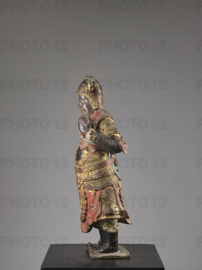 Lokapala — Guardian of the World; creator, Chinese Buddhism; figurine sculpture of a warrior in polychrome red, brown, black; Qing dynasty 1644–1911; 19th century ; full sculpture; height 72.5 cm; China; material: sculpture., A painted and gilded standing figure of a warrior shown in profile, wearing an ornate helmet and layered armor with intricate decorative patterns; the figure has a contemplative facial expression with arms positioned in front, and a draped skirt-like lower garment with visible folds. The visible colors include gold, red, black, brown, and traces of green, with worn areas showing a darker underlayer. The figure stands on a small rectangular base placed on a flat black plinth against a neutral gray background.