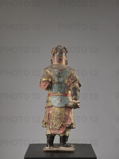 Lokapala Guardian of the World, creator unknown, sculptor; polychrome wooden full sculpture of a warrior figurine in red, brown, black; Chinese Buddhism; Qing dynasty 1644–1911, 19th century, China; height 72.5 cm., A painted and gilded carved wooden figure shown from the back, standing on a rectangular base; the figure wears layered armor and draped garments with ornate raised patterns, including a textured central panel on the lower back, and holds an object in one hand; visible colors include muted blue, faded red, gold, black, green, and areas of exposed wood and brown; the figure has a helmet with decorative elements and detailed sleeves and boots, and the background is a smooth neutral gray.