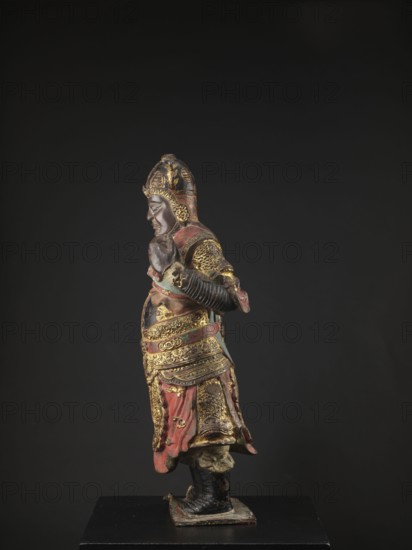 Lokapala — Guardian of the World; unknown sculptor; Chinese; Chinese Buddhism; figurine of a warrior; Qing dynasty 1644–1911; full sculpture; polychrome red, brown, black; 19th century ; China; sculpture; height 72.5 cm, A standing carved figure in profile wearing ornate armor and a helmet, with visible textures of carved metalwork and fabric; the figure has a contemplative facial expression and clasped hands at chest level, a layered skirt-like lower garment and high boots, and stands on a small rectangular base. The visible colors include black, gold, red, brown, and hints of green.