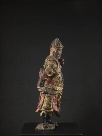 Lokapala — Guardian of the World; sculptor, polychrome full sculpture figurine of a warrior; Chinese Buddhism; Qing dynasty 1644–1911, 19th century, China; materials/polychrome colors: red, brown, black; height 72.5 cm., A painted sculpture of a standing figure shown in profile wearing ornate armor and a helmet, with visible carved facial features and a beard; the figure's attire includes layered decorative plates, a skirt-like lower garment, and a sash, with textured patterns and raised details on the armor and clothing. The statue displays colors of red, gold, black, and hints of green, with areas of worn paint and a slightly glossy finish on some surfaces. The figure stands on a small base that rests on a black plinth, and the background is a solid dark gray to black.
