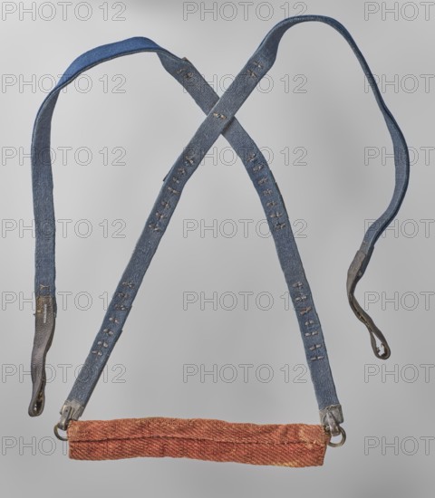 Tailpiece, repossession Salzburg; Chinese art; Chinese culture; Qing dynasty 1644–1911; riding equipment for horses; cloisonné enamel; fish motif; made of leather ribbon and bronze decorated with cellular enamel on copper colorful, on a blue background; 18th century; China; horse tack element; length 110 cm, A suspended textile harness with crossed straps forming an X shape, made of a textured blue fabric with visible stitched slits along the length of the straps and darker worn end tabs, attached at the bottom to a horizontal padded band in a textured orange-red fabric with metal rings and fittings; the background is a smooth light gray.