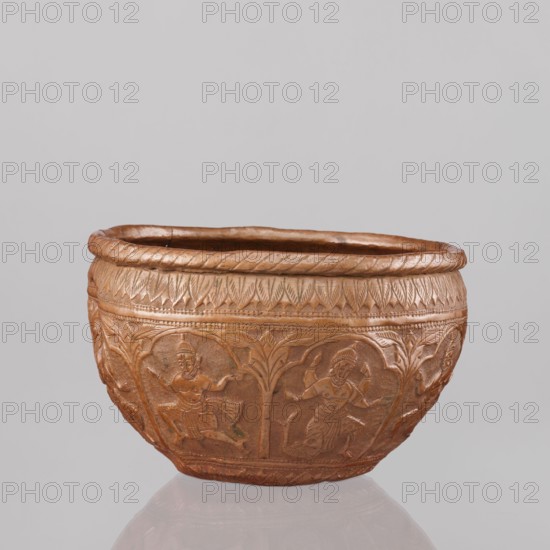Bowl, Vishnu depicted; figures of the god Vishnu placed in arcade poses. Decorated with avatars, turtles, plant ornaments, geometric ornaments, lions, fish, wild boars, flood scenes, trees. At the top and bottom an ornament of stylized palm leaves. 19th century, India. Vessel. Height 11.5 cm, diameter 18.1 cm., A rounded decorative bowl with a warm brown and coppery surface sits against a pale gray background, its exterior covered in carved reliefs of human figures in dynamic poses framed by stylized plant motifs, with a braided rim and bands of repeating leaf-like patterns encircling the upper portion, and subtle darker brown shading in recessed areas that emphasizes the carved details.