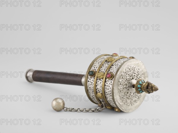 Prayer wheel, Tibetan, 19th century. Cult object used in Buddhist rituals; scrolls inside. Materials not specified. Height 28 cm., An ornate handheld object lies on a pale gray surface against a light gray background, featuring a dark brown cylindrical handle with silver-toned end caps attached to a decorative cylindrical head. The head combines silver-toned filigree with gold-toned bands and raised gold-toned accents, and is embellished with small rounded stones in green, pink, and turquoise colors set into the bands and a protruding finial. A short silver-toned chain with a slightly tarnished round silver-toned weight is attached to the head.