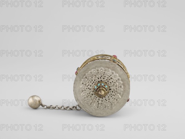 Prayer wheel, Tibetan, 19th century. Cult object used in Buddhist rituals; scrolls inside. Height 28 cm., Materials not specified., A round metallic object with ornate filigree work on its visible face rests on a light background, showing a central raised knob decorated with small turquoise-colored accents and surrounded by intricate pierced floral patterns; the outer rim appears silvery-gray and slightly worn, while the edge and some adornments display a warm gold tone with a few pinkish-red and blue-green beads set around it; a short chain of interlocking links extends from the object to a spherical weight that shares the silvery-gray color and shows slight tarnish.