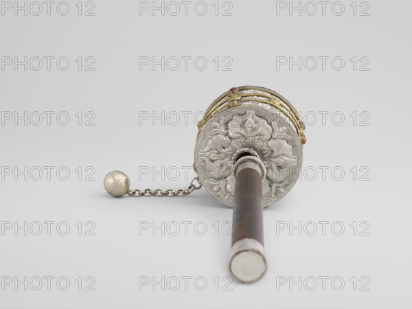 Prayer wheel, Tibetan, 19th century. Cult object used in Buddhist rituals; scrolls. Materials not specified. Height 28 cm., An object resembling a cylindrical handheld device lies on a pale background, showing a circular ornate end with raised floral patterns in a silver tone, a wooden shaft in a dark brown color extending from the center, and a connected chain with a metal spherical weight in a dull silver color; the circular end also features several narrow bands in a gold color with small colored beads in red and turquoise attached.