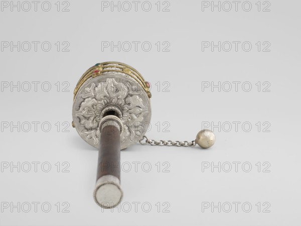 Prayer wheel;, Tibetan Buddhist ritual cult object; scrolls; 19th century; Tibet; height 28 cm., An object photographed against a pale gray background showing a cylindrical wooden handle extending toward the viewer attached to a round ornate metal head with a raised floral motif; the head has decorative metallic bands and small round colored stones in green and red set into the bands, and a short metal chain with a spherical metal weight lies to the right of the head; visible colors include silver, gold, brown, green, red, and pale gray.
