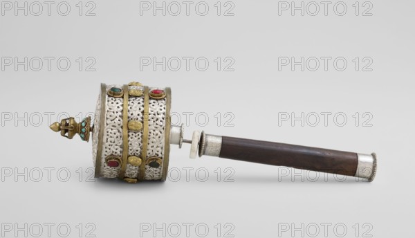 Prayer wheel, repossession, Tibetan Buddhist art, Tibetan culture, Buddhist ritual scrolls, 19th century, Tibet, cult object, height 28 cm, A handheld cylindrical object with a dark brown wooden handle and silver metal end cap lies horizontally on a light gray background; the cylinder head is ornate with white filigree panels framed by gold bands and embellished with several round colored stones in red, green, turquoise, and dark tones, and a small gold finial with turquoise accents projects from one end.