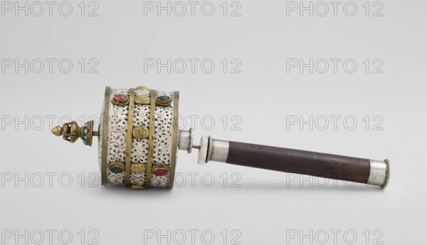 Prayer wheel, Tibetan Buddhist ritual scroll/cult object, 19th century, Tibet; height 28 cm., An ornate cylindrical object with a long dark brown wooden handle lies horizontally against a light gray background; the cylinder features an intricate white filigree pattern accented by vertical gold bands and gold decorative round elements, with several small colorful stones in shades of red, green, and turquoise set into the decorative bands; the left end of the cylinder has a gold finial with turquoise accents and a pointed tip, while the right end connects to the wooden handle through silver-toned fittings with a beaded rim, and the overall visible colors include white, gold, dark brown, silver, red, green, turquoise, and light gray.