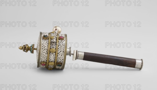 Prayer wheel, Tibetan Buddhist ritual scroll/cult object, 19th century, Tibet, Tibetan culture, Buddhist art, height 28 cm., A handheld object lies horizontally against a pale gray background, consisting of a cylindrical drum-like section attached to a dark brown wooden handle with silver-colored metal fittings; the drum section shows ornate openwork metal with filigree patterns in a silvery color, accented by gold-colored bands and decorative gold-colored elements, and set with several small colored stones in shades of blue, turquoise, and pink; one end of the drum has a pointed finial in a brass or gold tone with turquoise-colored inlay, and a small silver-colored spherical bead is visible near the top; the handle is dark brown wood with silvery metal collars at both ends.