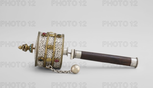 Prayer wheel, Tibetan Buddhist ritual scroll/cult object, 19th century, Tibet; height 28 cm; Tibetan Buddhist art., An ornate handheld object rests on a light gray background, featuring a cylindrical decorative drum section attached to a dark wooden handle. The drum section displays intricate openwork metal filigree in a silvery color with several horizontal brass bands and round bezel-set stones in colors including red and green. A small brass finial with turquoise-colored insets projects from one end of the drum, and a short metal chain with a spherical weight in a dull gold color hangs from the drum. The wooden handle is dark brown with silvery metal fittings at the joint and at the end. The overall color palette includes silver, brass/gold tones, dark brown, red, green, turquoise, and light gray.