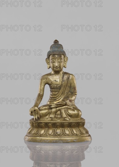 Buddha sitting on a lotus throne;, ; figurine; Chinese culture; Chinese art; Buddha iconography; Buddhist art; lotus throne iconography; ushnisha; Qing dynasty 1644–1911; 19th century; possibly China/Tibet; sculpture; height 14.4 cm., A gilded statue of a seated figure on a rounded lotus base, shown against a plain light gray background. The figure sits cross-legged with one hand resting on the knee and the other in the lap, wearing a draped robe with decorative patterns and a textured topknot. The surface shows areas of gold and darker patina, with touches of blue on the hair and subtle darker spots across the body and base. The base is carved with repeating petal motifs and the statue casts a faint reflection on the surface below.
