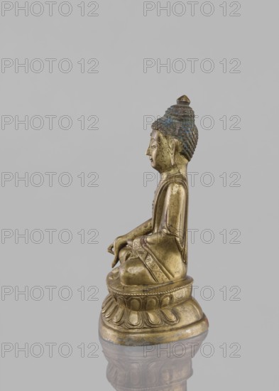 Buddha sitting on a lotus throne;, ; figurine; Chinese culture; Chinese art; Buddha iconography; Buddhist art; lotus throne iconography; Harbin, China; Qing dynasty 1644–1911; ushnisha; 19th century; possible Tibetan origin; sculpture; height 14.4 cm., A small metallic statue of a seated figure shown in profile on a rounded pedestal with a carved lotus motif, with a reflective surface beneath creating a faint mirrored reflection; the figure has a textured hair or headdress with traces of blue-green coloration and a pointed top, a serene facial expression, elongated earlobes, and a robe draped over the shoulders with decorative patterns; visible colors include gold, blue-green, and gray for the background and reflection.