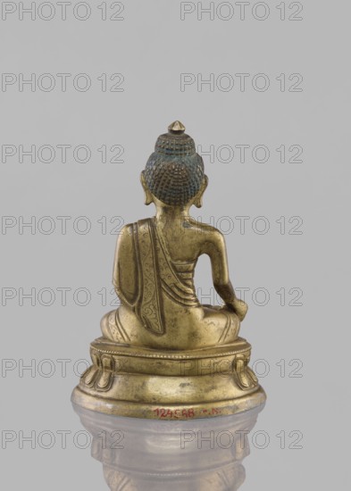 Buddha sitting on a lotus throne, figurine, Chinese culture, Chinese art, Buddha iconography, Buddhist art, lotus throne iconography, Harbin, Qing 1644–1911, ushnisha, 19th century, Tibet, sculpture, height 14.4 cm, A small metallic statue of a seated figure shown from the back on a rounded base, with a detailed textured hairstyle topped by a small pointed element and visible blue-green patina on the head; the figure wears a draped garment with engraved decorative patterns and sits with one arm resting on the knee, all rendered in a warm golden-bronze color, set against a neutral gray background and reflected faintly on a glossy surface below.